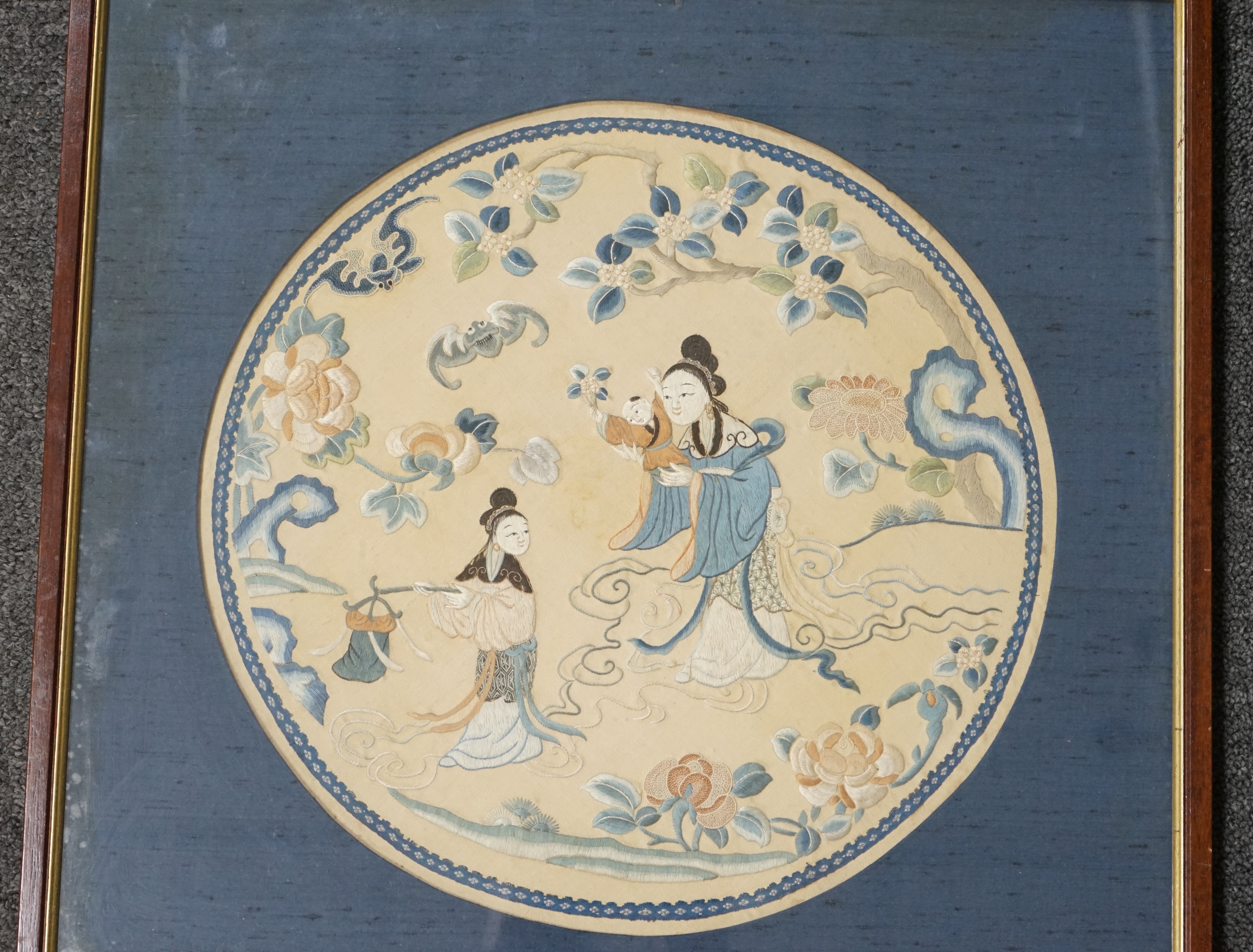 A Chinese embroidered silk roundel, 19th century, depicting immortals on swirling clouds amid blossom, flowering shrubs and a bat, the central female immortal holding a child, in raised silk and Chinese not on a pale pea